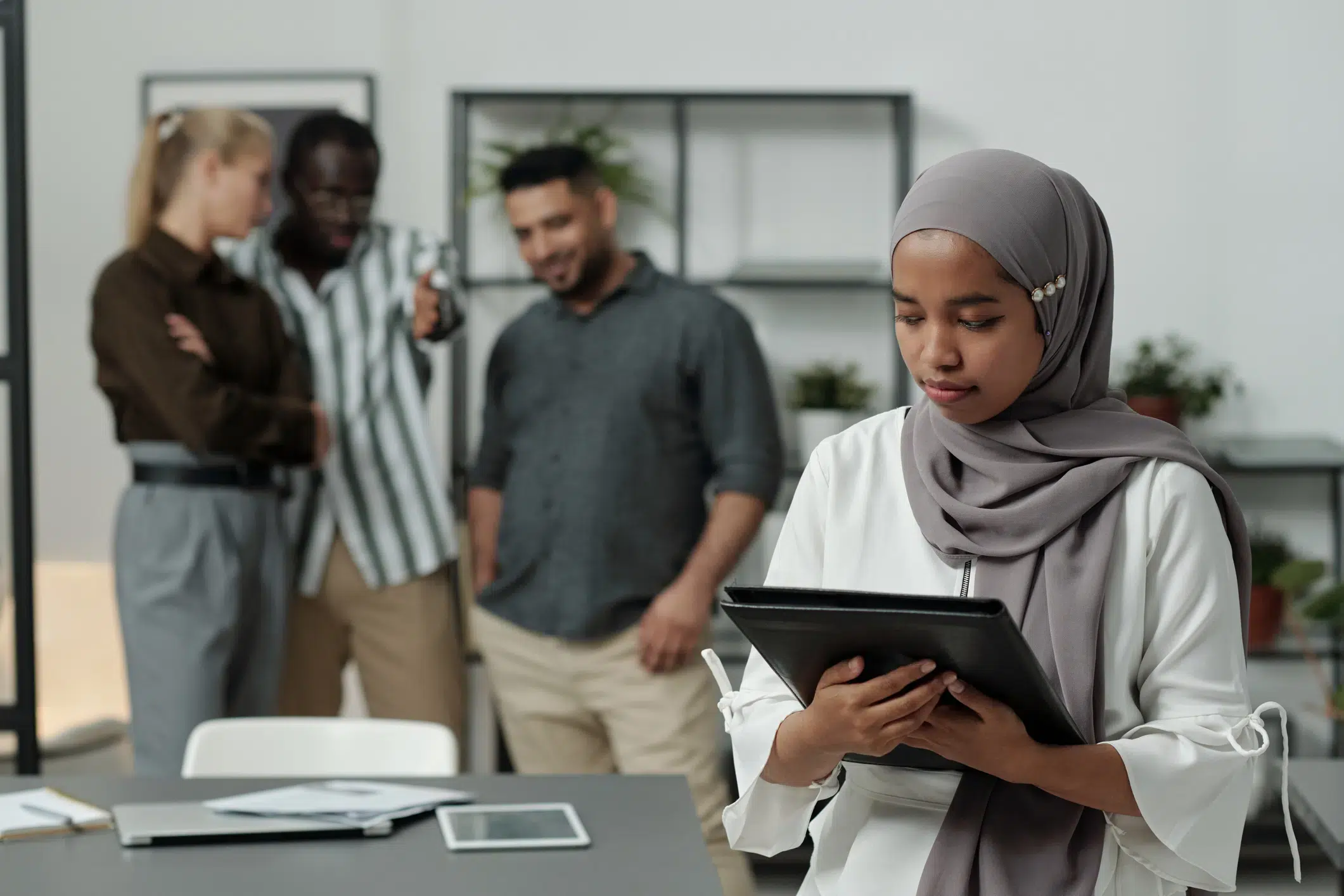 Young Muslim woman in a hijab using a tablet while two men in the background laugh, suggesting workplace exclusion or racial bias.